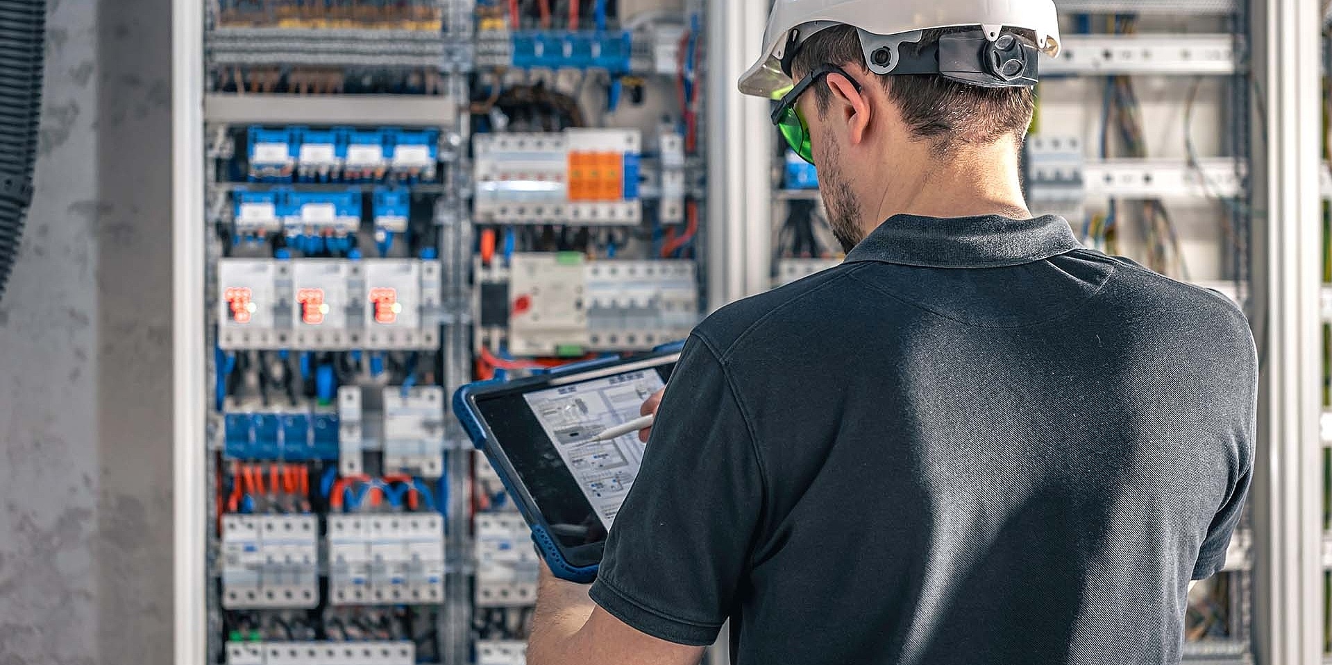 Man, an electrical working in a switchboard with fuses, uses a tablet.